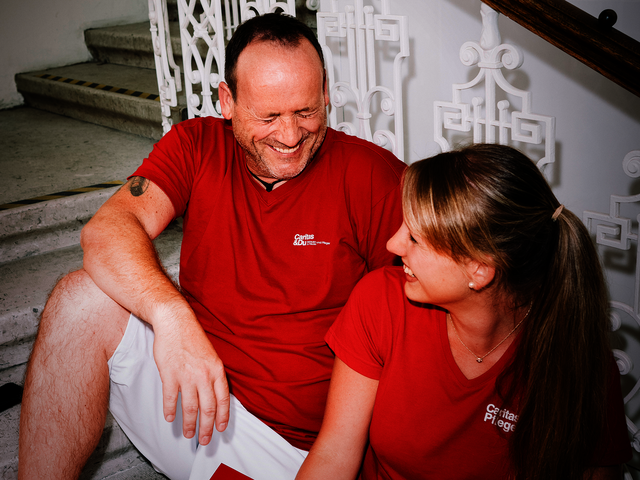 Ein Mann und eine Frau mit roten T-Shirts mit Caritas-Logo sitzen auf den Stiegen und lachen gemeinsam. Die Frau hat einen Aktenordner in der Hand.