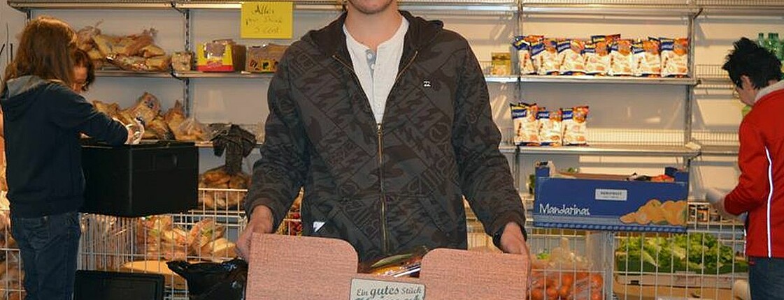 Ein junger Mann steht in einem Solidarmarkt und hält eine Kiste voller Lebensmittel in der Hand.