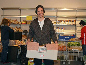 Ein junger Mann steht in einem Solidarmarkt und hält eine Kiste voller Lebensmittel in der Hand.