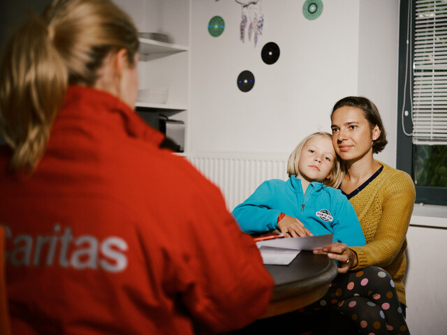 Eine Mutter sitzt mit ihrem Kind auf dem Schoß. Ihnen gegenüber am Tisch sitzt eine Frau mit einer roten Caritas-Jacke, sie ist von hinten zu sehen. 