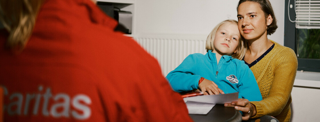 Eine Mutter sitzt mit ihrem Kind auf dem Schoß. Ihnen gegenüber am Tisch sitzt eine Frau mit einer roten Caritas-Jacke, sie ist von hinten zu sehen. 